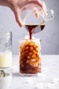 A hand pouring cold brew coffee into a glass filled with ice cubes.