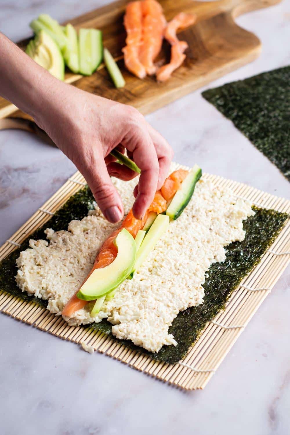 A hand placing avocado on a piece of salmon that is on a bed of cauliflower rice spread across nori on a bamboo sheet.