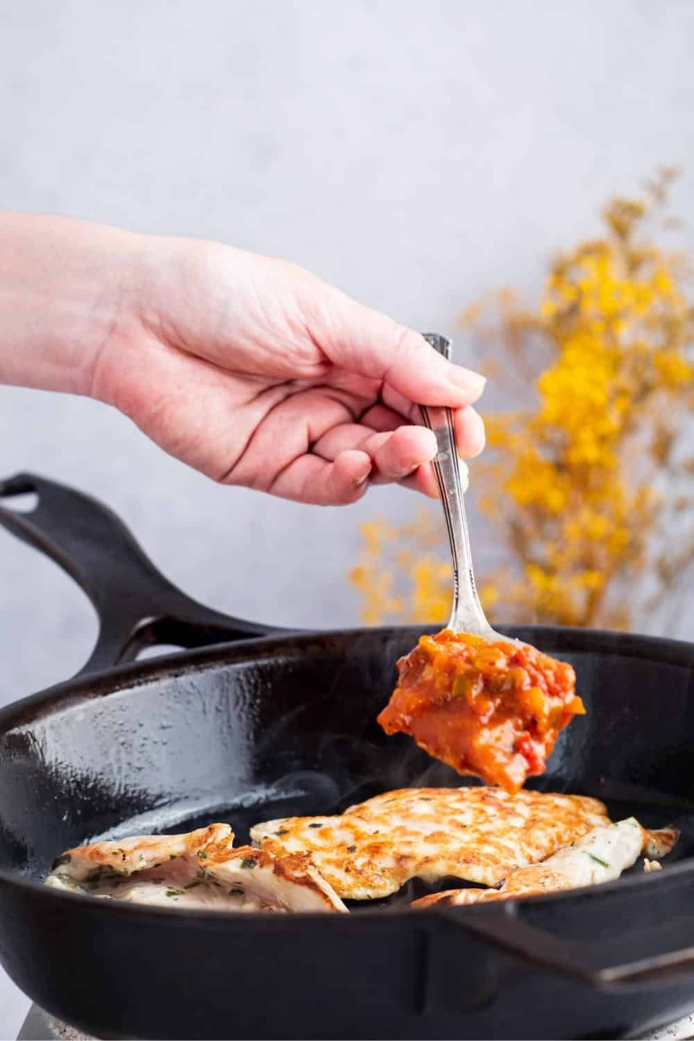 A hand pouring sauce on top of a piece of grilled chicken in a skillet with two more pieces of grilled chicken in it.