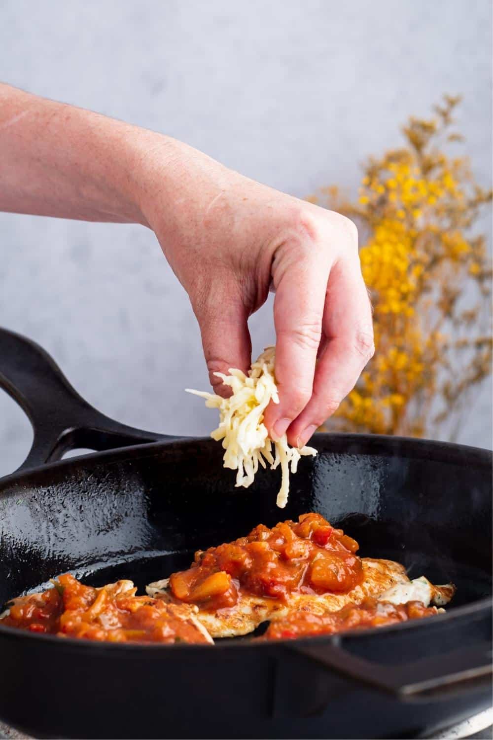 A hand holding shredded cheese over three pieces of chicken with sauce on top in a black skillet.