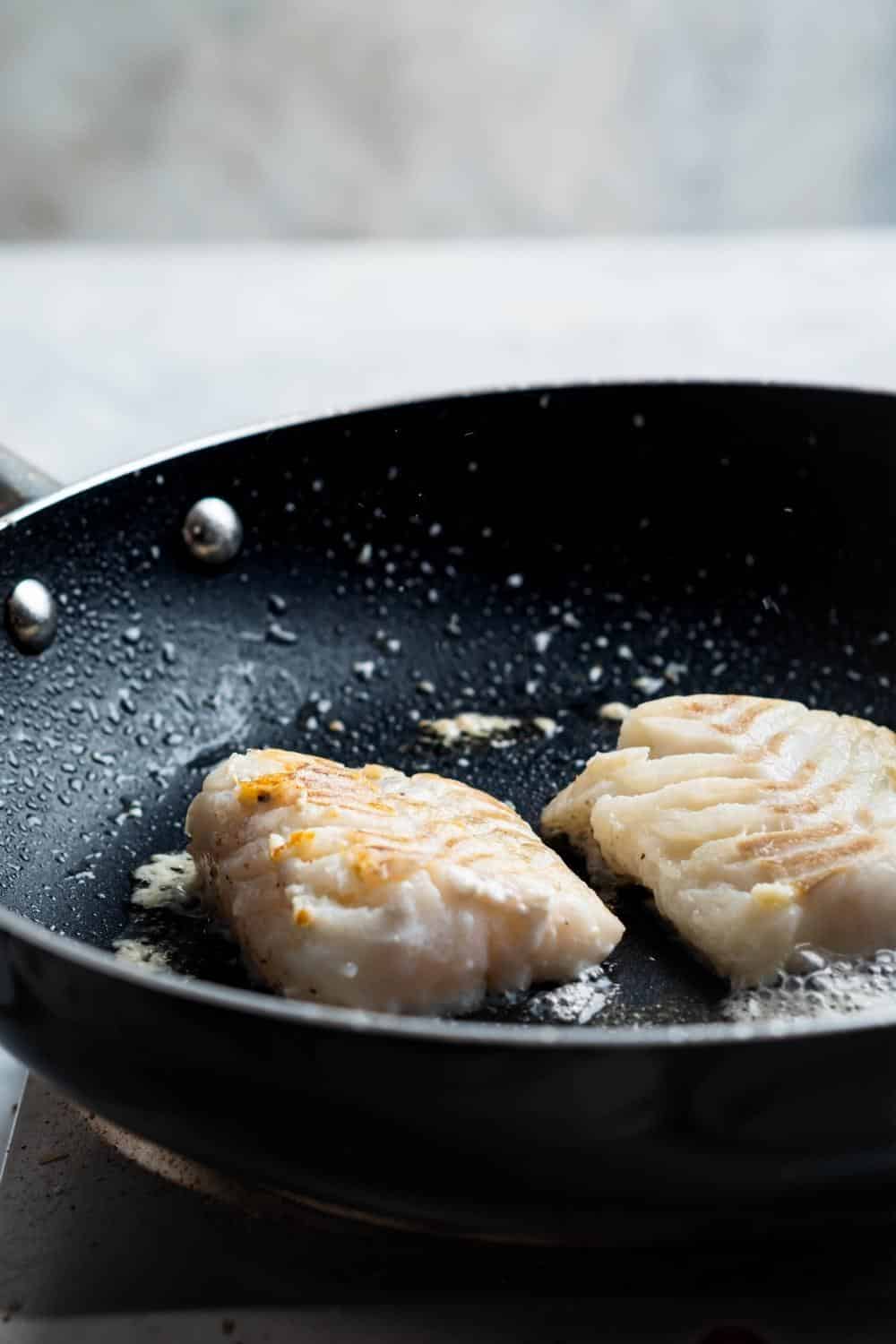 Two halibut fillets cooking in a black skillet.