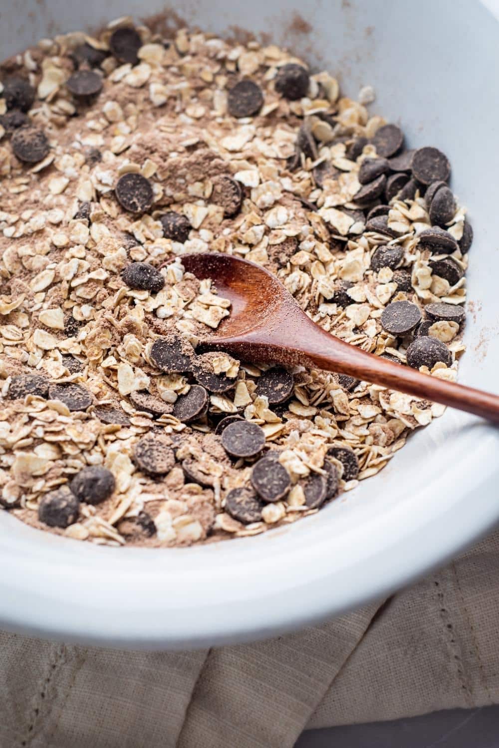 A wooden spoon in a bowl with oats, chocolate protein, and chocolate chips.