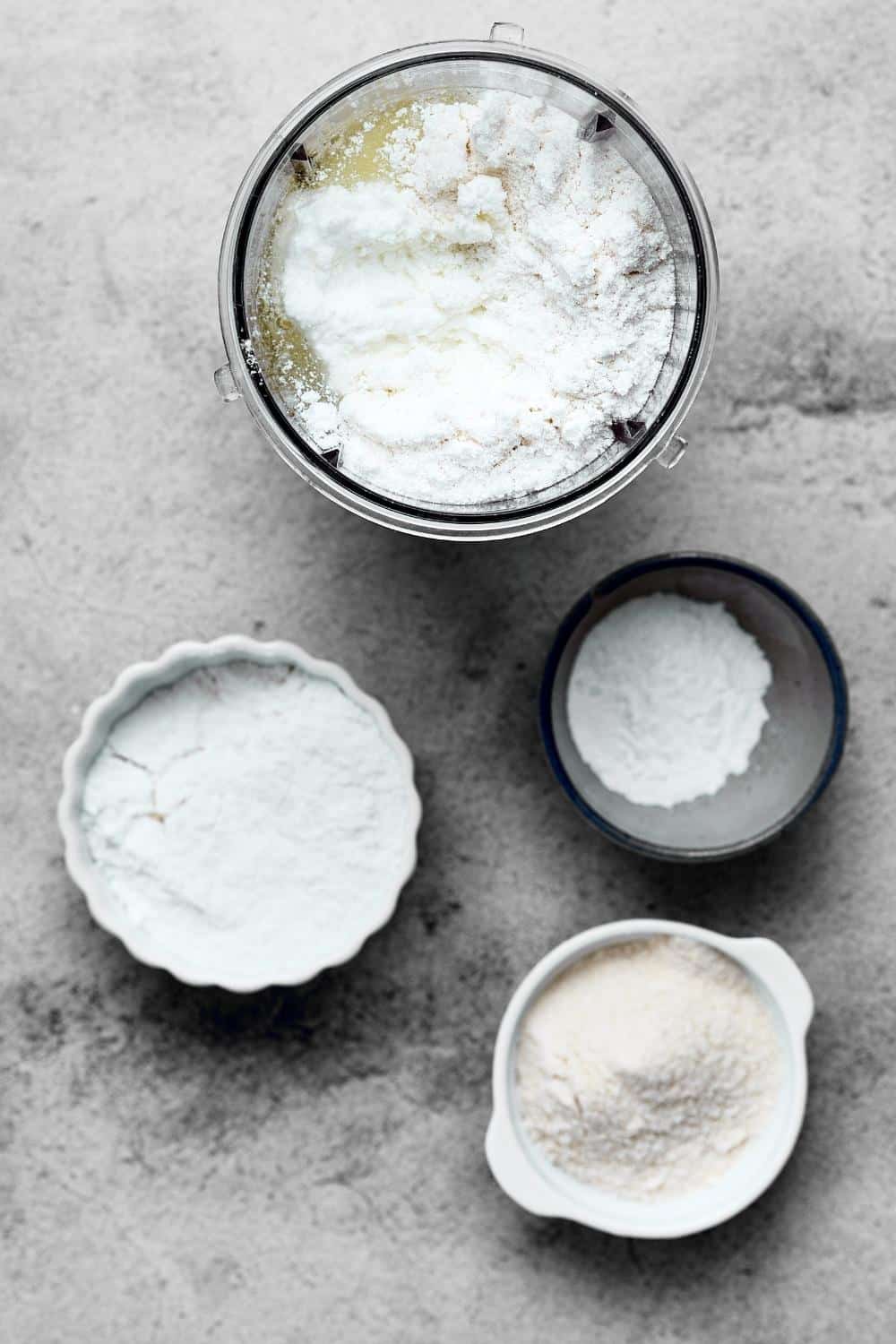 A blender with pancake ingredients in it, a bowl of vanilla protein powder, and a bowl of sweetener on a gray counter.