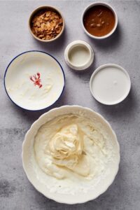 A bowl of mixed heavy cream and coconut milk, a bowl of erythritol, a bowl of almond butter, and a bowl of toasted almonds all on a grey counter.