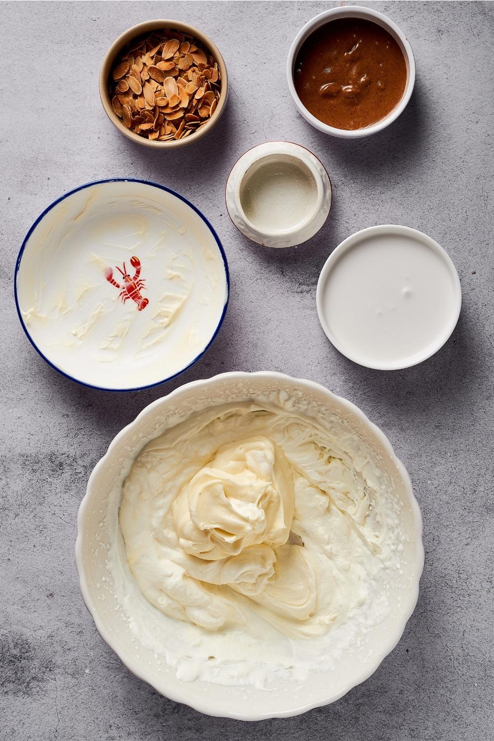 A bowl of mixed heavy cream and coconut milk, a bowl of erythritol, a bowl of almond butter, and a bowl of toasted almonds all on a grey counter.