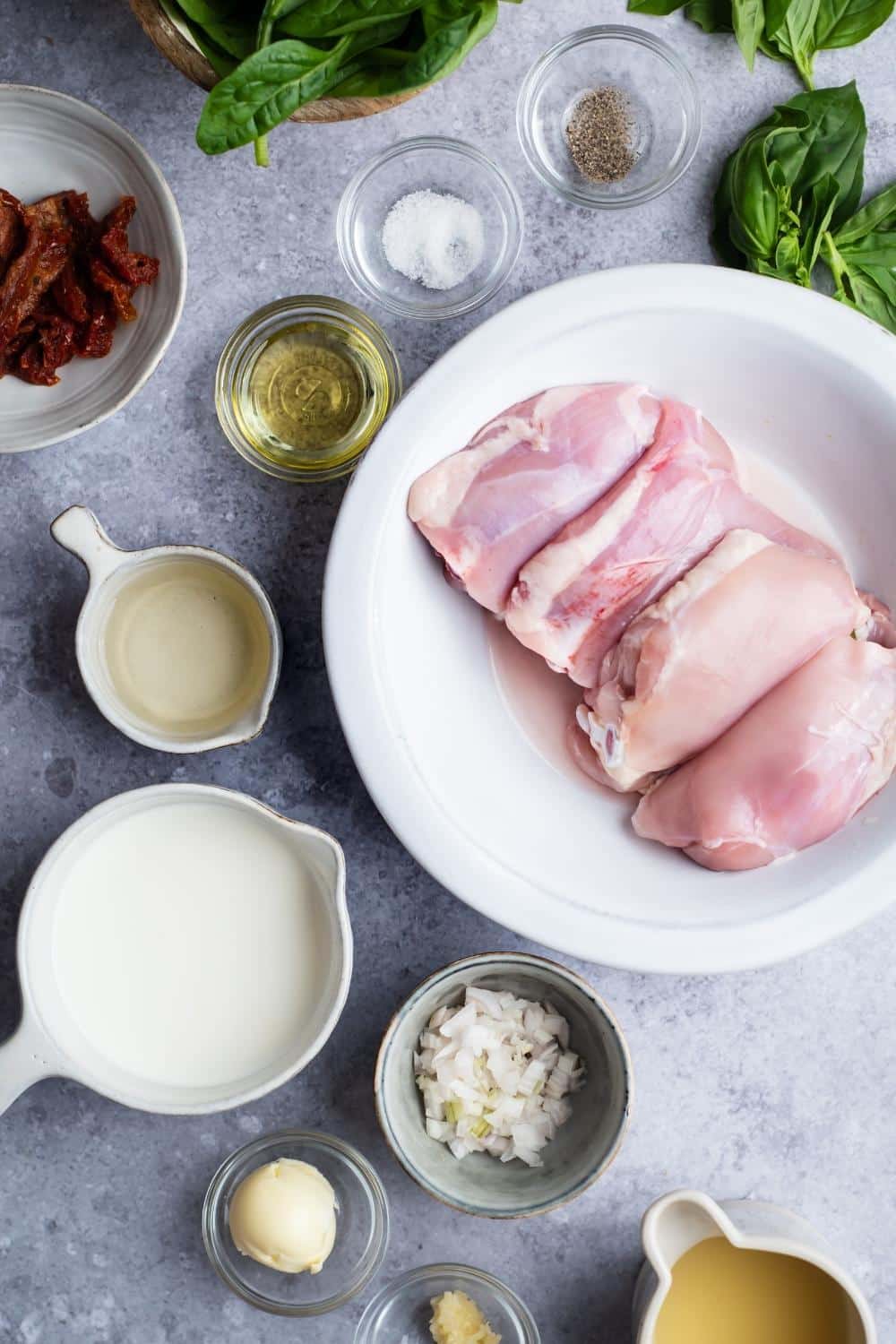 Chicken thighs on a plate, diced onions in a bowl, cream sauce in a bowl, olive oil in a bowl, salt and pepper in bowls all on a grey counter.