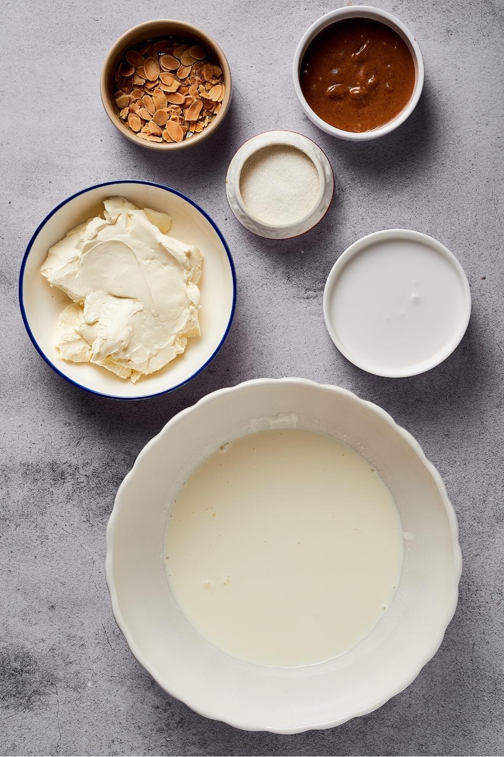 A bowl of coconut milk, a bowl of almond butter, a bowl of erythritol, a bowl of cream cheese, and a bowl of heavy cream all on a grey counter.