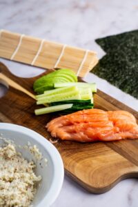 A wooden cutting board with salmon and sliced cucumber on it.
