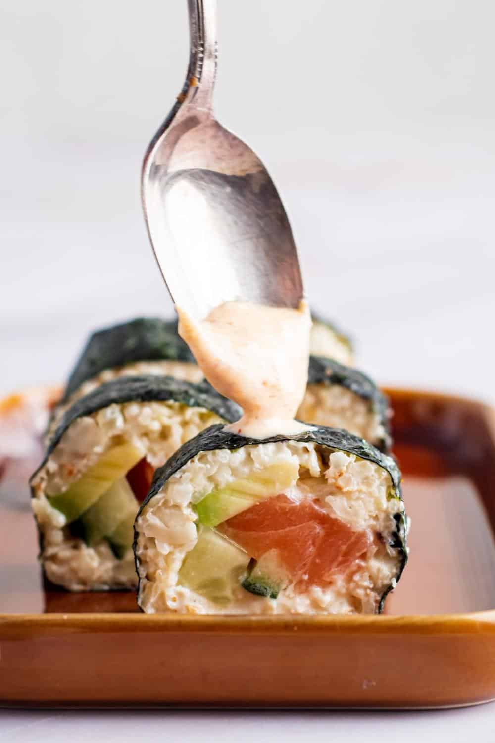 A spoon drizzling sriracha mayo on top of a keto sushi roll. There are some more keto sushi pieces behind it.