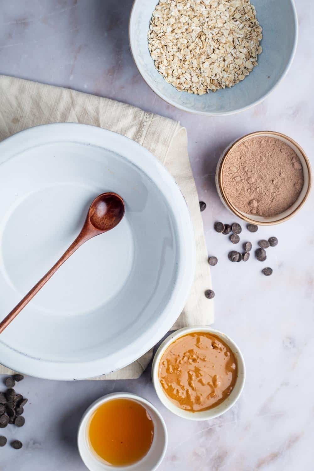 A bowl of honey, a bowl of peanut butter, a bowl of protein powder, and a bowl of oats all on a white counter.