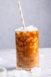 A glass with a straw and ice cubes in it filled with protein coffee. The glass is on a white counter and there are some ice cubes in front of the glass.