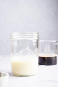 A glass jar partially filled with milk. Behind it is a glass cup partially filled with iced coffee.