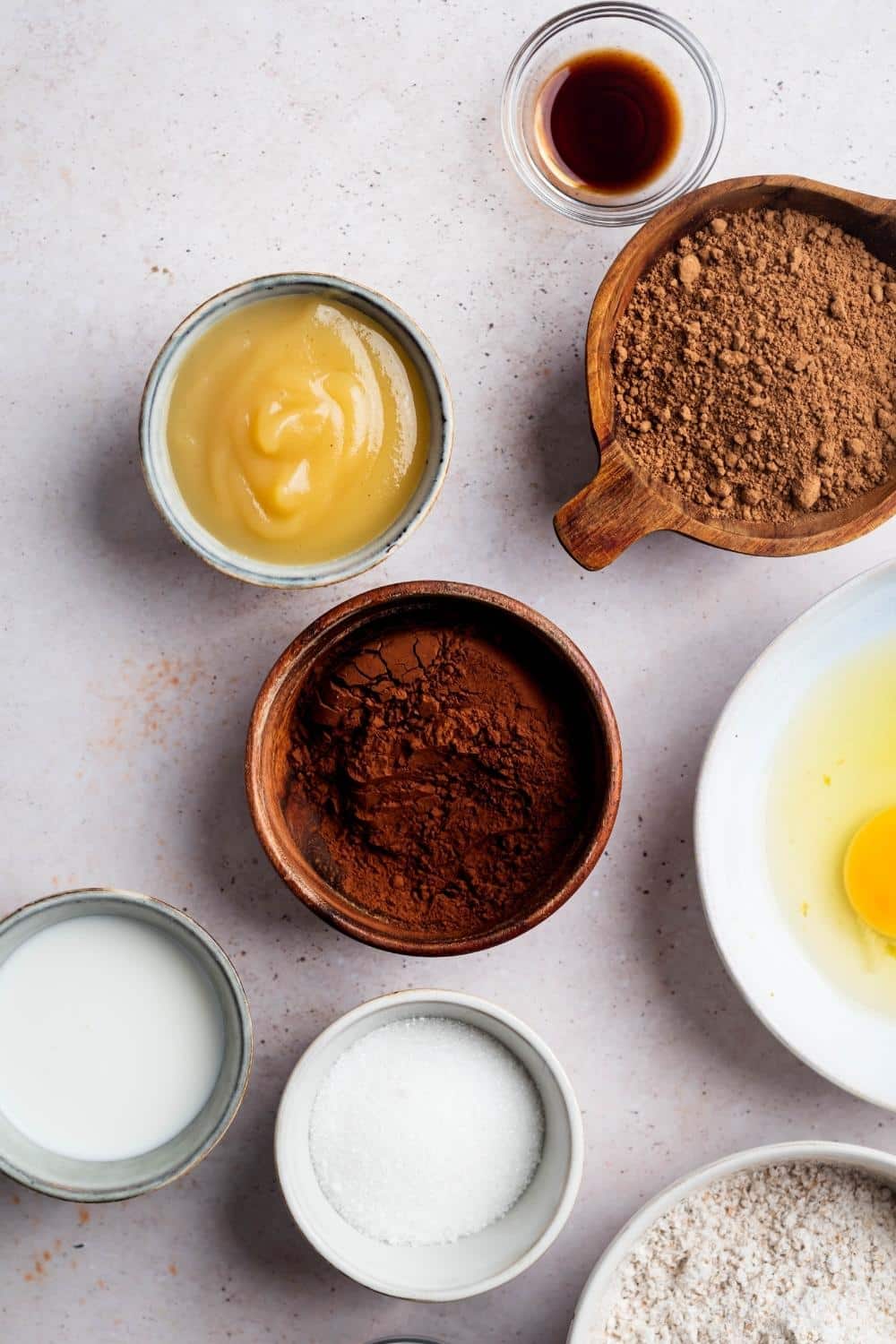 A bowl of sugar substitute, a bowl of milk, a bowl of cocoa powder, a bowl of applesauce, a bowl of chocolate protein powder, and a bowl of vanilla extract all on a white counter.