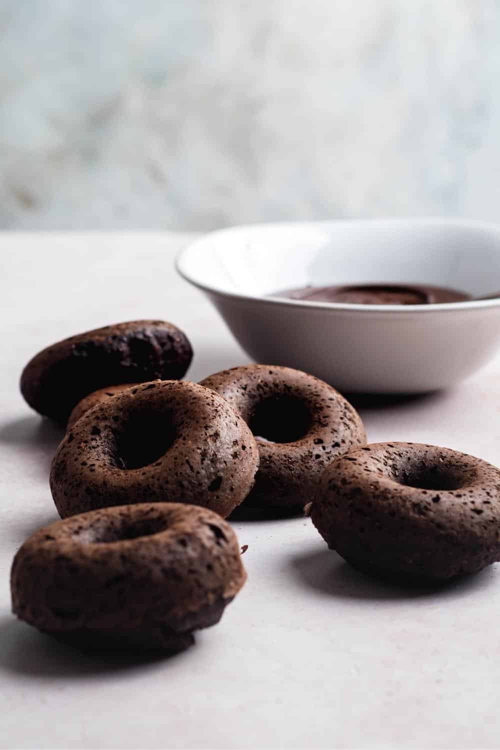 Five chocolate protein donuts on a white counter with a white bowl filled with chocolate glaze behind them.