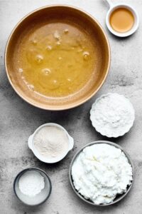 A bowl of egg whites, a bowl of vanilla protein powder, a bowl of sweetener, a bowl of cottage cheese, and a bowl of baking powder all on a white counter.