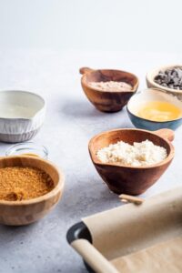 A bowl of coconut sugar, a bowl of almond flour, a bowl of butter, and a bowl of chocolate protein powder on a white counter.