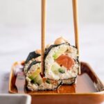 Two chopsticks with a piece of sushi between them being held over a serving dish.