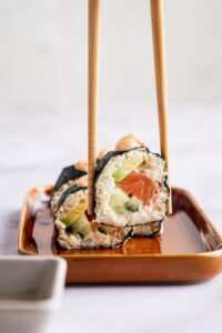 Two chopsticks with a piece of sushi between them being held over a serving dish.