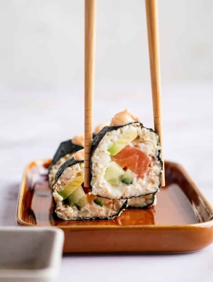 Two chopsticks with a piece of sushi between them being held over a serving dish.