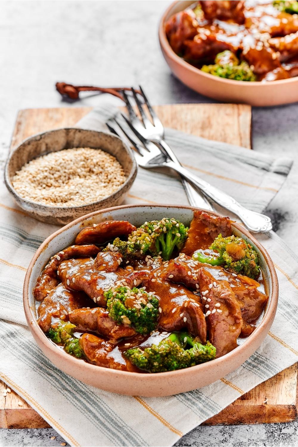 A bowl of beef and broccoli stir fry on a tablecloth with a bowl of rice behind it.