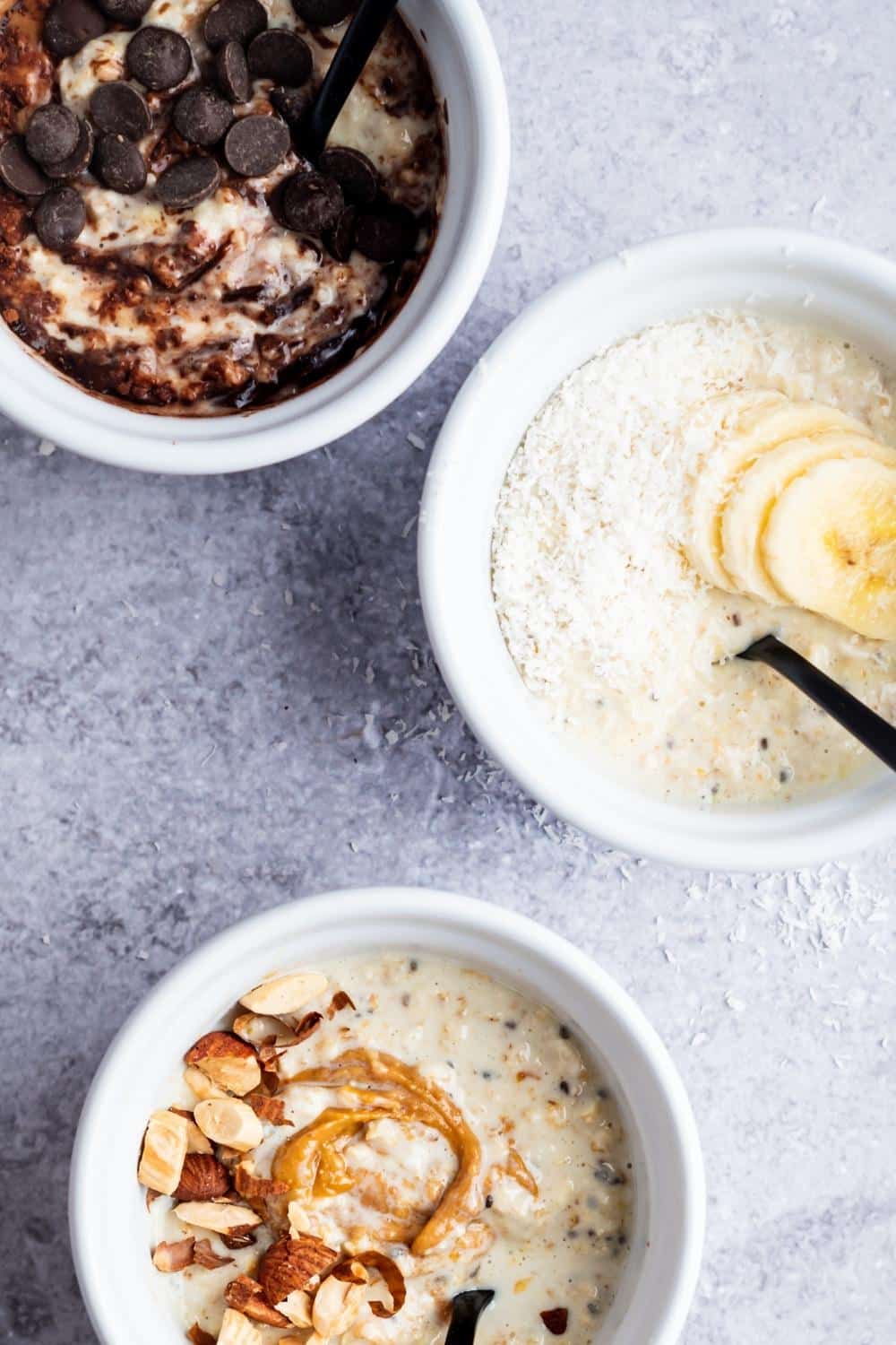 Half a bowl with peanut butter and nuts on top of overnight oats, part of a bowl with sliced bananas and shredded coconut, and part of a bowl with chocolate chips and cocoa powder on top of overnight oats.