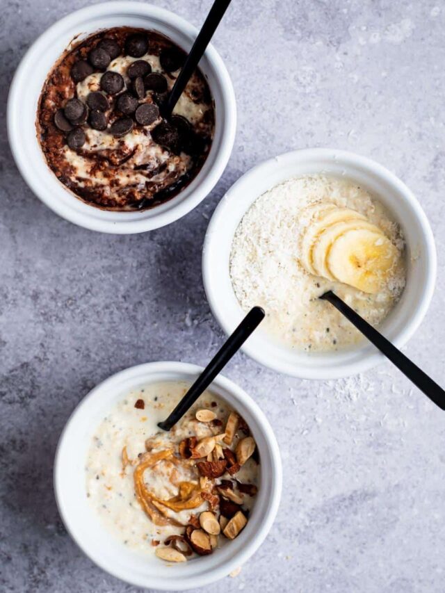 Chocolate chips and cocoa powder on top of overnight oats in a white bowl, banana slices and shredded coconut on top of overnight oats in a white bowl, and nuts and peanut butter on top of overnight oats in a white bowl. All of the bowls are in a line on a grey counter.