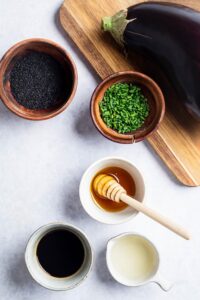 A bowl of honey, a bowl of diced green onions, a bowl of sesame oil, and part of an eggplant on a cutting board.