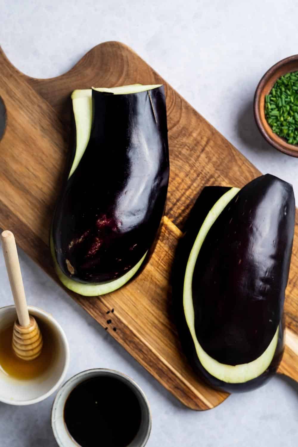 An eggplant cut in half and sliced lengthwise on a wooden cutting board.