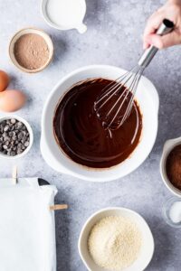 A hand whisking brownie batter in a bowl with a bowl of chocolate chips, a bowl of almond flour, and a bowl of cocoa powder surrounding it.