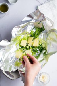 A hand putting greens and ginger root on a sheet of tin foil in a casserole dish.