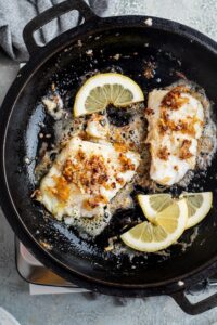 Two mahi mahi fillets cooking in a skillet with lemon wedges.