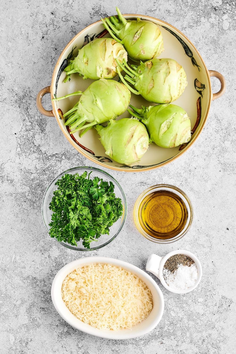 A bowl with kohlrabi in it, parlsey in a bowl, oil in a bowl, salt and pepper in a bowl, and grated parmesan in a bowl.