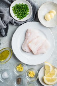 Two mahi mahi fillets on a plate, a bowl of garlic, a bowl of lemon, a plate with butter, and a plate with lemon wedges all on a grey counter.