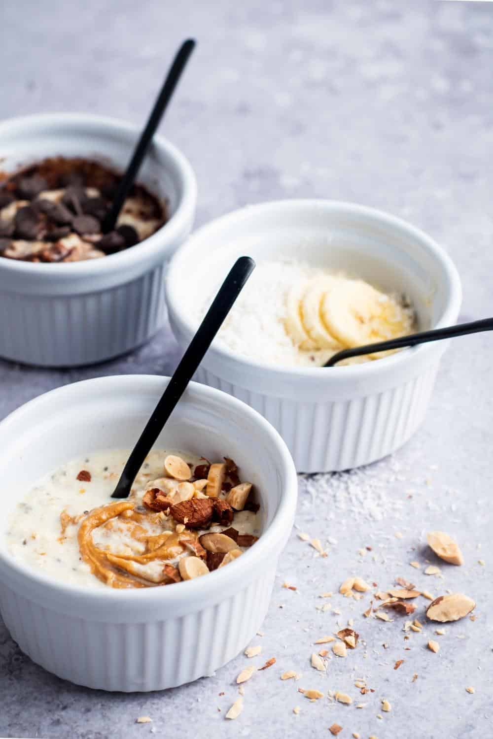 Nuts and peanut butter on top of overnight oats in a white bowl. Behind that is sliced bananas and shredded coconut in a white bowl. And behind that is another bowl of overnight oats