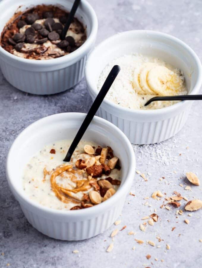 A bowl of protein overnight oats that has peanut butter and nuts on top of it, behind that is another bowl of protein overnight oats with sliced banana and coconut slices on it, and behind that is a bowl of protein overnight oats with cocoa powder and chocolate chips on top.