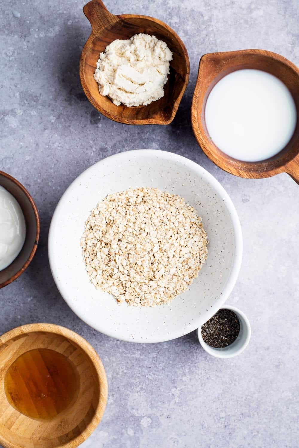 Rolled oats in a bowl, protein powder in a bowl, milk in a bowl, Chia seeds in a small bowl, and honey and wooden bowl on a gray counter.