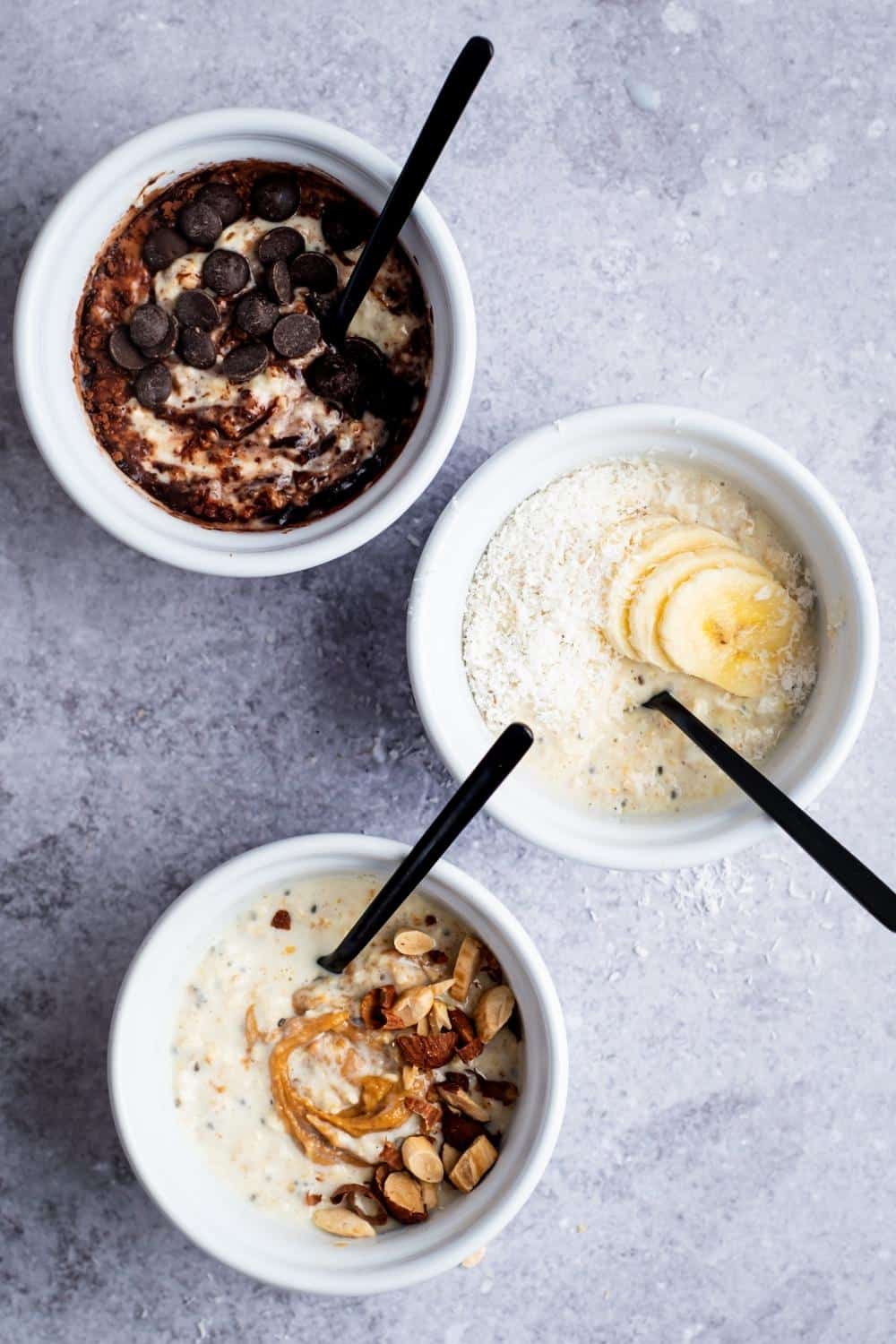 Chocolate chips and cocoa powder on top of overnight oats in a white bowl, banana slices and shredded coconut on top of overnight oats in a white bowl, and nuts and peanut butter on top of overnight oats in a white bowl. All of the bowls are in a line on a grey counter.