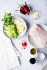 A whole sea bream on a plate, a bowl of soy sauce, a bowl of sesame oil, greens and cheese on a plate, and a bowl of chili peppers all on a grey counter.