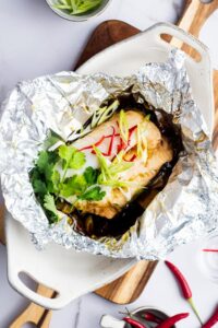 Cooked sea bream in tin foil in a white casserole dish on a wooden cutting board.