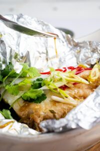 A spoon drizzling sauce on top of a whole sea bream in tin foil.