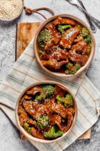 Two bowls filled with beef and broccoli stir fry on a tablecloth on a cutting board on a grey counter.