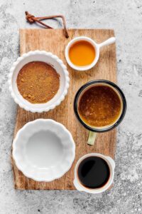 A bowl of egg yolk, a bowl of brown sugar, a bowl of soy sauce, an empty on a wooden cutting board on a grey counter.