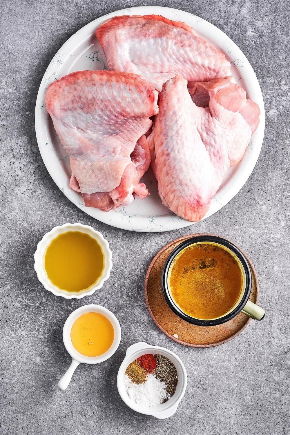 Three raw turkey wings on a white plate. In front of it a bowl of honey, a bowl of seasonings, and a bowl of stock.