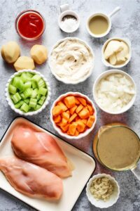 Two chicken breasts on a plate, sliced carrots, sliced celery in a bowl, cream cheese in a bowl, chopped onion in a bowl, hot sauce in a bowl, and potatoes all on a grey counter.