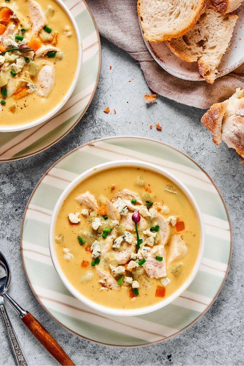 Buffalo chicken soup in a white bowl on top of a stripped plate on a grey counter. Behind it is part of another bowl of soup and some bread.