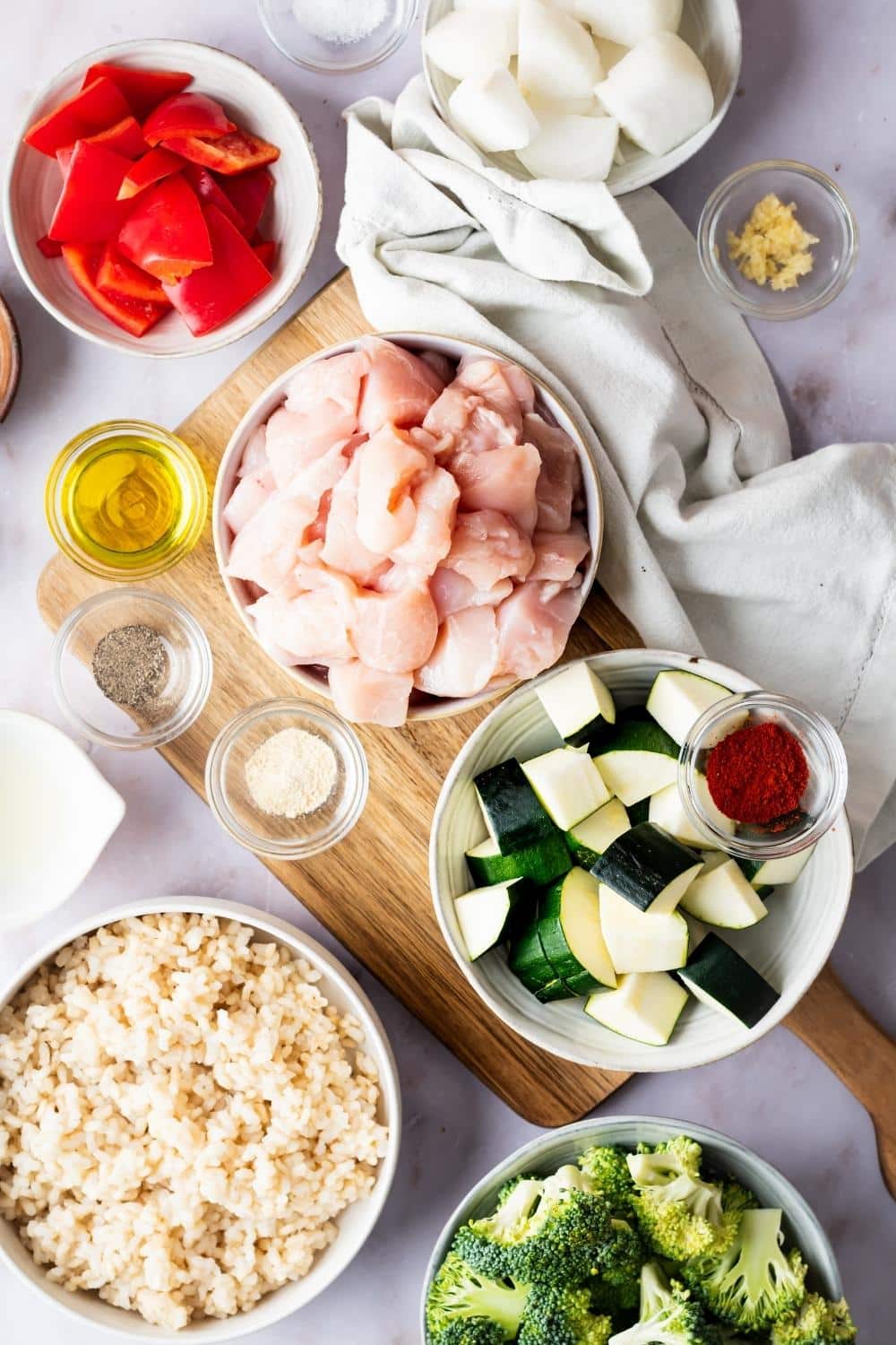 A bowl of chicken, a bowl of zucchini, a bowl of rice, a bowl of red bell pepper, a cup of oil, a cup of salt, a cup of pepper, and part of a bowl of broccoli all on a counter.