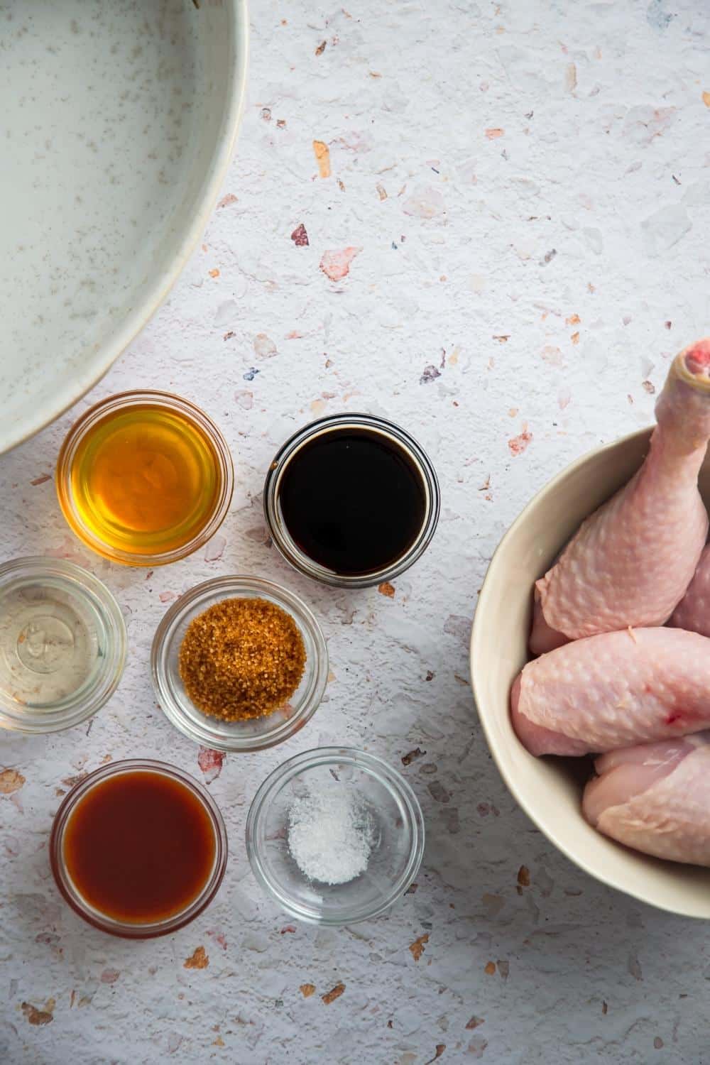 A bowl of soy sauce, a bowl of honey, a bowl of brown sugar, a bowl of rice vinegar, and part of a bowl with chicken legs in it.