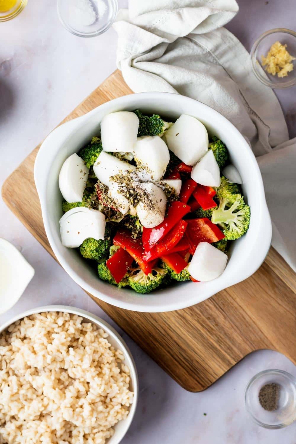 Onion, broccoli, and red bell pepper in a bowl on a wood cutting board. In front of that is part of a bowl of rice.