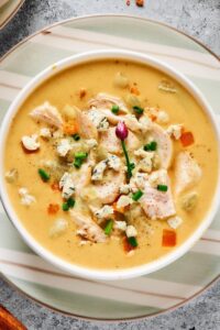 Buffalo chicken soup in a white bowl on part of a stripped plate on a grey counter.