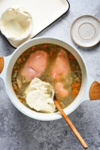 Cream cheese and chicken breasts in a bowl of broth and diced veggies.